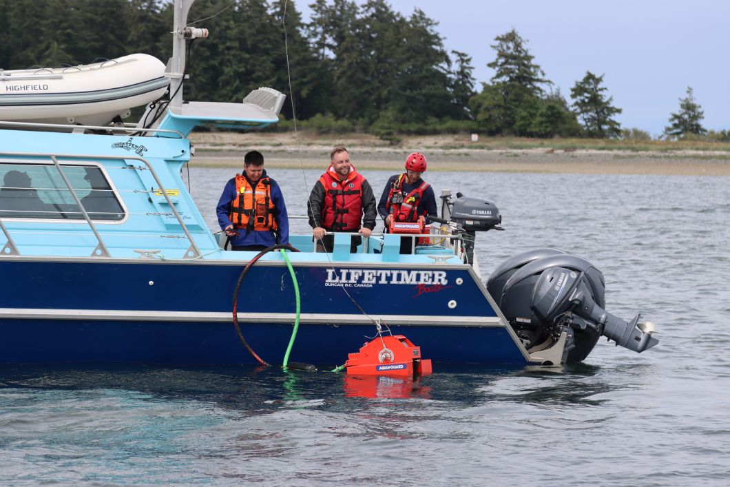 Three people stand on the side of a boat as they operate a skimmer. The skimmer is red and in the shape of a large box similar to a lawn mower. 